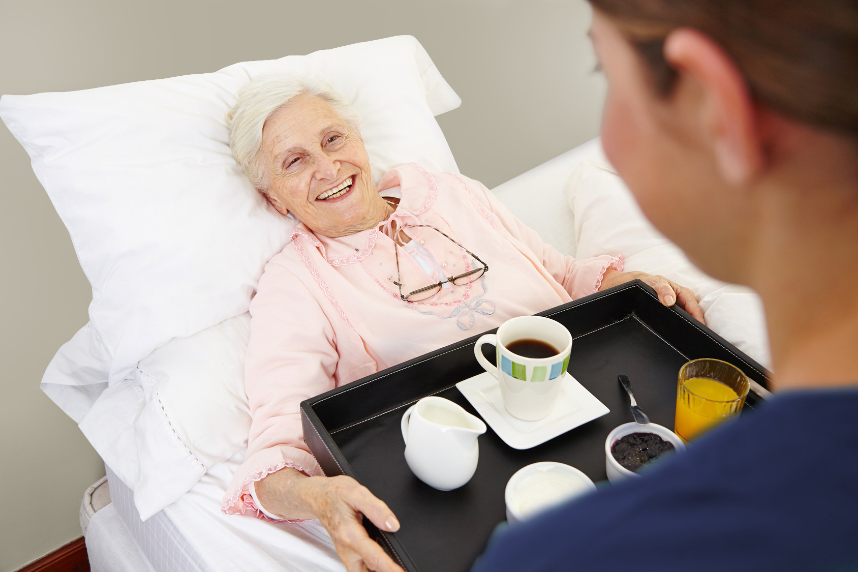 Eine alte Frau die im Bett liegt und von einer Pflegerin ein Tablett mit Kaffee gereicht bekommt.